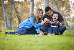 Couple with a baby sitting in a grassy field blowing bubbles and smiling.