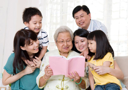 Large family of 5 huddling around an elderly woman reading from a pink book.